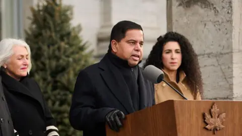 Getty Images Public Safety Minister Gary Anandasangaree stands outside before a podium and a microphone, wearing a black coat, sweater and gloves. He is flanked by two women. 