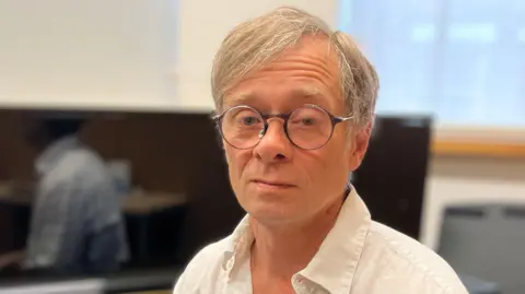 Alex Dunlop/BBC Alex Street has short blonde hair. He is wearing black framed glasses and a white shirt. He is sitting next to a piano which is in a room next to a window.