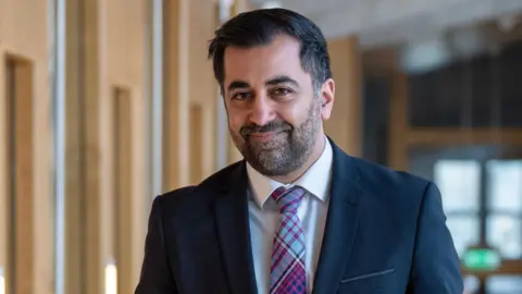 Humza Yousaf wearing a dark suit jacket, white shirt and patterned tie standing in a brightly lit indoor corridor with wooden-panelled walls and large windows in the background