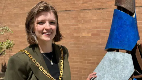 Gavin Kermack / BBC A young woman with neck-length mid-brown hair wearing a dark green blazer over a black top. She has a chunky gold chain around her neck and is resting her hand on the artwork from before.