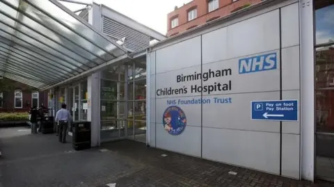 The Birmingham Children's Hospital trust sign is visible by the entrance to the hospital with glass roofing and frontage. It is black and blue writing, with a circular picture with an animated child in its centre.