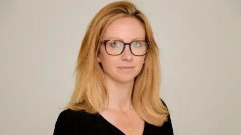 Lucy Rigby in an official portrait image. She is dressed in black, wearing tortoiseshell-patterned glasses and a gold pendant necklace. She has shoulder length hair