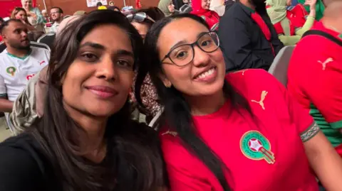Manal Two young women smile at the camera. One is wearing a Moroccan football jersey, the other is wearing a black t-shirt.