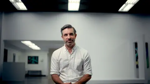Brandon Silverman, a man with dark hair with a touch of grey, a side-parting and stubble, wearing a white shirt and pictured sitting down in what appears to be a large gallery space with mostly bare white walls and bright overhead lighting.