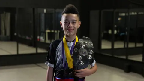 A boy in a mirrored studio smiles at the camera holding a trophy and wearing a belt that reads Oireachtas Rince na Gaeilge. He has tight curls cut into a mohawk style