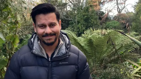 José Palma smiles at the camera, he is wearing a black puffer jacket and has a goatee and moustache. There are plants behind him.