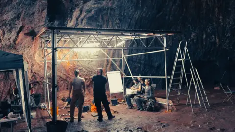 A small scaffolding structure with fluorescent lights casting light onto a small dug out area with people working in it. The structure sits within a dark cave with brown and green textured walls. Ladders, buckets and other equipment can be seen among people looking on.