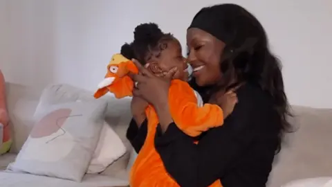 A young baby wearing an orange onesie is held up to her mother's face as the two exchange smiles