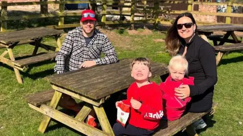Andrew and Zoe Family photograph of dad Andrew, mum Zoë, and their sons Joey, who is four, and his little brother, two-year-old Tommy. It is a sunny day and they are sitting around a wooden picnic bench