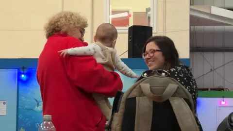 A woman wearing a red jacket holds a baby wearing beige dungarees. A second woman wearing glasses and a black dress with white spots smiles at the baby.