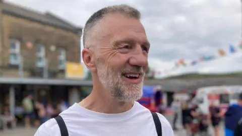 A man with greying short hair and a beard stands laughing in the street.