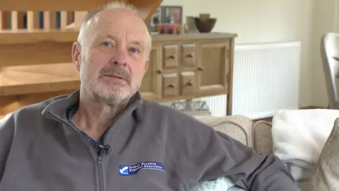 A man wearing a grey jumper is sitting on a sofa inside a house. There is wooden furniture behind him. 