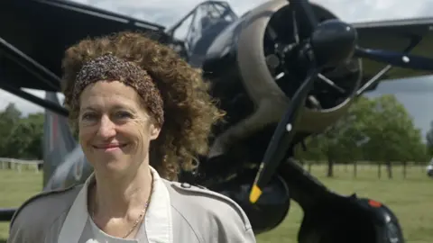 Jamie Niblock/BBC A woman with thick, brown curly hair and and bandana holding her hair back stands in front of a World War Two aircraft. She is smiling and wears a grey coat.