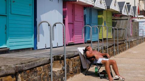A man sunbathes in front of a row of colourful beach huts in Walton-On-The-Naze, Essex