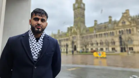 Aisha Iqbal/BBC A young man of South Asian descent stands in a city square. Behind him out of focus is a large, classical town hall building with a clock tower. He wears a black coat and a black and white scarf.