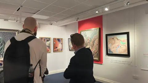 Looking from behind at two people looking at the artwork in part of the gallery space. Paintings go around all the walls, which are mostly white with one red section. All paintings are surrealist with vibrant colours, unusual shapes and curves. All very neatly and precisely painted.