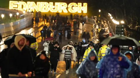 PA Media People walking in the rain in Edinburgh on Hogmanay in 2022. There is an illuminated Edinburgh sign in the background. The street is busy with people holding umbrellas or with hoods or jackets over their heads.