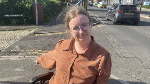 An image of Alicia Cartwright, she is wearing an orange blouse with blue diamonds and is seated in her wheelchair. She is pointing at a section of pavement she says is dangerous.