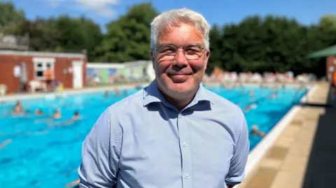 A man with grey hair and glasses is standing by an outdoor swimming pool. People can be seen in the water behind him