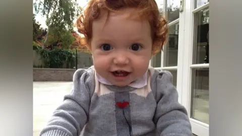 Team Margot A child with curly red hair smiles at the camera while sitting in a garden. She is wearing a gray sweater with red hearts