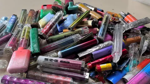A pile of disposable vapes collected by Laura Young is tipped out onto a table.