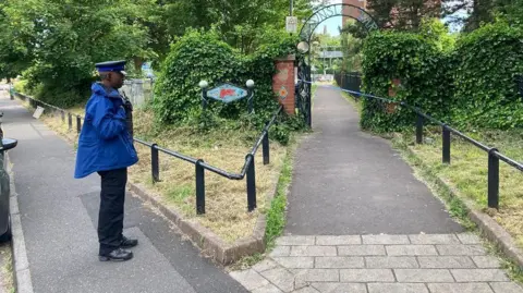 Entrance to a park - with wrought iron archway - cordoned off, with officer keeping watch