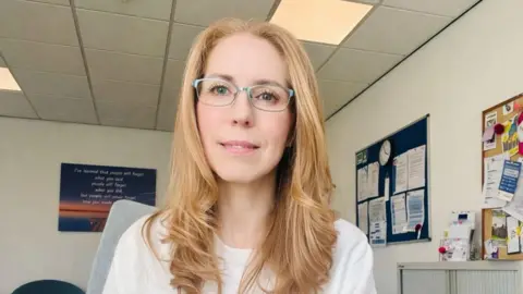 Zoe Bostock has long blonde hair and glasses, and wears a white top. She is pictured standing in what appears to be a surgery, smiling slightly.