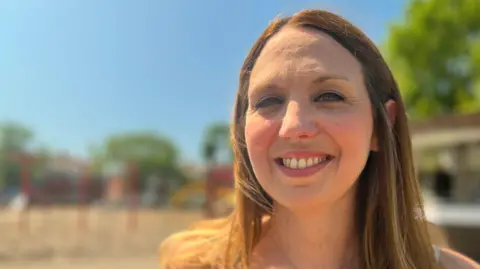 A picture of Sophie from the neck up looking into the lens. She has brown-blonde hair. The background is out of focus but a children's play park can be made out.