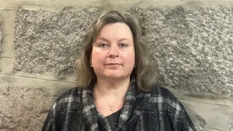 Claire Moggach, a woman with shoulder-length, dark blonde hair wearing a black, grey and white top. She is photographed in front of a granite wall.
