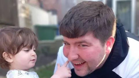 A female toddler with dark hair pointing her finger at the nose of a man leaning forward towards her