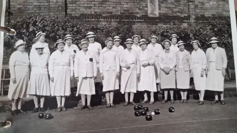 Contributed A black and white image of a group of women on a bowling green. They wear matching cardigans and skirts, with black shoes and bowler hats. Black bowling balls rest on the green in front of them.