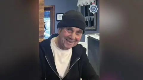 John McColl wearing a black hat and white t-shirt, with a black hoodie, is sitting indoors. 