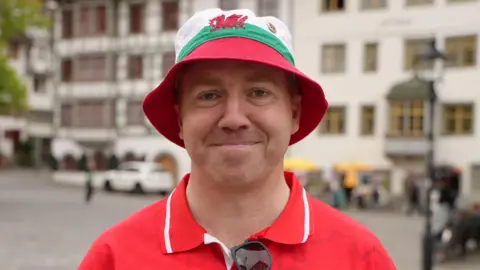 Gareth Reid smiling at the camera. He is wearing a red and white Wales shirt and a red, white and green bucket hat with the Welsh dragon at the front. He is standing on a street with yellow cars behind him. 
