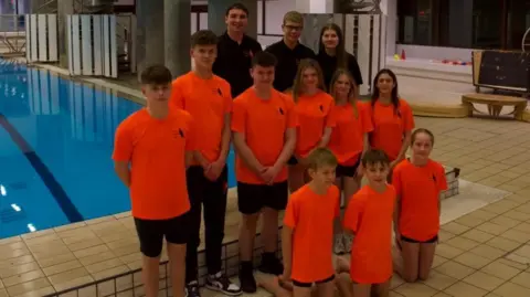 Ayr Diving Club A group of kids wearing orange t-shirts stand in front of two spring boards over a bright blue swimming pool 