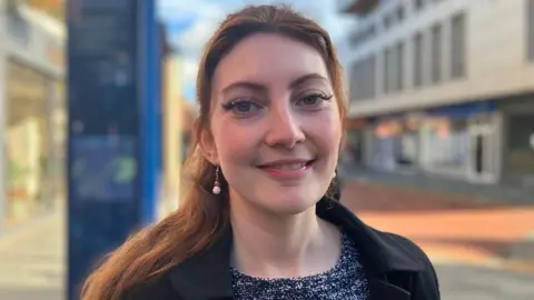 A head-and-shoulders photo of Anna Davies, a woman with ginger hair and wearing a black coat, smiling at the camera with a parade of shops behind her.