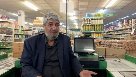 Rehman sits at a till in his shop - behind him you can see the cash register and aisles of grocery items