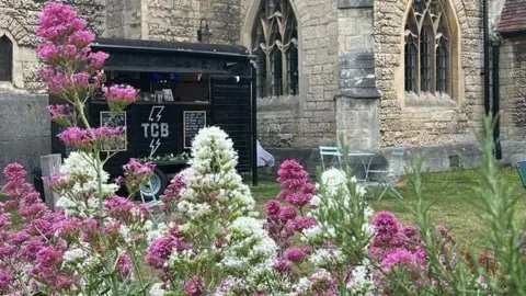A coffee van, which is black, outside a church and on grass. In front of the van are purple and white flowers.