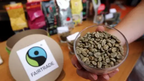 Getty Images A hand holds a glass bowl of coffee beans towards the camera. In the background, a lid with the fairtade logo can be seen in front of bags of coffee.