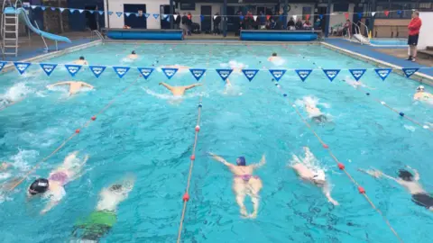 Swimmers in Stanhope Open Air Swimming Pool. Strings of bunting hang widthways over the pool and a lifeguard stands on the side of the pool. Lanes are marked out with ropes and buoys and the approximately 14 swimmers in shot all appear to be doing the butterfly stroke. At the far end of the pool a number of people sit or stand in front of a white building which has doors and windows with dark frames. On the left hand side of the pool stands a slide, much like a family with a child might have in their garden. A silver ladder leads to the top but the blue slide winds towards the pool.