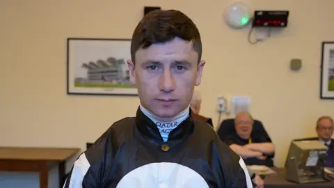 Qays Najm/BBC Jockey Oisin Murphy looks directly at the camera slightly smiling. He has short dark hair and wears a jockey uniform. Jeremy Lind can be seen sitting at a desk behind him. 