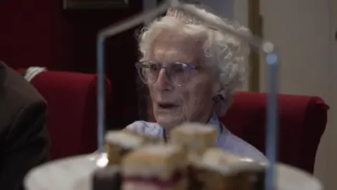 Cilla sits in a restaurant. A plate of cakes is in the foreground.