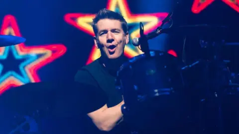 Getty Images Dougie Vipond playing the drums. he is looking at the camera with his mouth open, like he is singing. He is wearing a dark T-shirt and the drums are in the dark but his face is lit up by a spotlight. There are coloured stars behind him.