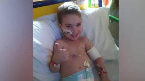 Family photo Finley as a young boy, lying in a hospital bed with tubes attached to his body, arm and face. He is wearing a nappy. He is smiling at the camera and giving a thumbs-up with his right hand.