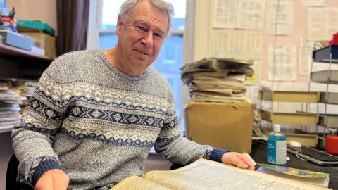 Ray smiles at the camera, wearing a grey fairisle-style jumper. He has open in front of him the yellowed pages of an old newspaper. Blurred behind him are stacked newspapers on a box and noticeboard.