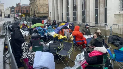 A crowd of people in warm clothing are gathered outside The Halls in Wolverhampton. Many are sat in collapsible chairs and some hold umbrellas.