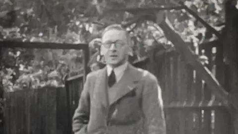 Black and white image of Cyril Tong. He is wearing a suit and tie and is stood in front of a garden fence