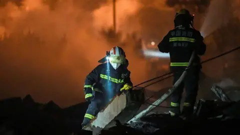Xinhua Firefighters walk around debris, with an orange haze behind them.