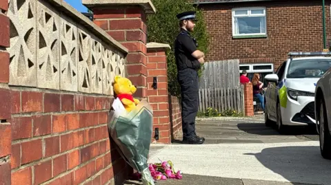 BBC A couple of bouquets of flowers leaning against a brick wall with a Winnie the Pooh teddy bear. There is a police officer dressed in black standing further down with his arms crossed. There are vehicles parked in front of them, including a white police car. A group of people can be seen gathered on the drive of an adjacent brick house. 