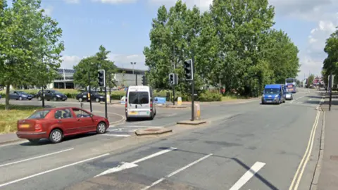 The road junction of Gloucester Road (A38) and Dorian Road with traffic lights and vehicles driving through, with Horfield Leisure Centre - a large grey building on the left and trees lining the roads in the background. 
