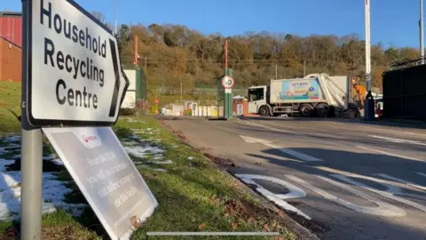 The entrance to a recycling centre with a white sign that reads "household recycling centre" which is on a grass verge.